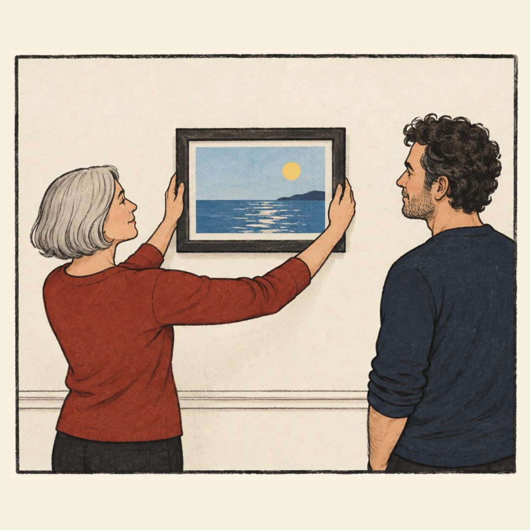 A silver-haired woman in a brick-red shirt holds both sides of a level framed seascape. A dark-haired man in navy watches. A horizontal molding crosses the ivory wall behind them.